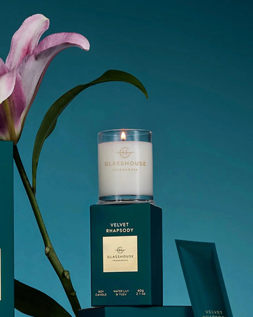 a white candle labeled "Glasshouse" on a green box, with a pink flower and a green leaf in the background.