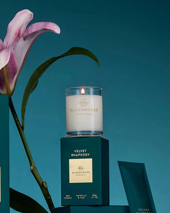 a white candle labeled "Glasshouse" on a green box, with a pink flower and a green leaf in the background.