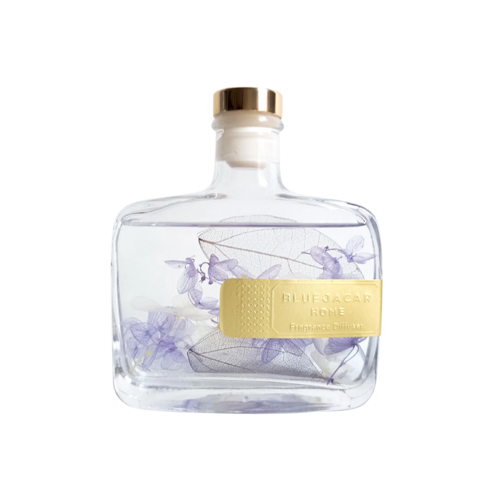 A clear glass bottle with a gold cap, containing a transparent liquid and decorated with purple flowers. The bottle has a yellow label that reads "BLUEOCAR HOME Fragrance Diffuser".