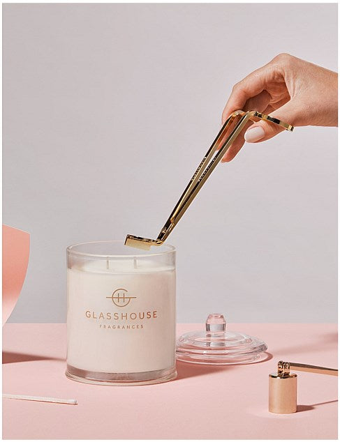A hand is holding a gold-colored candle holder, which is being used to light a white candle labeled "GLASSHOUSE Fragrances" on a pink background.