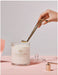 A hand is holding a gold-colored candle holder, which is being used to light a white candle labeled "GLASSHOUSE Fragrances" on a pink background.