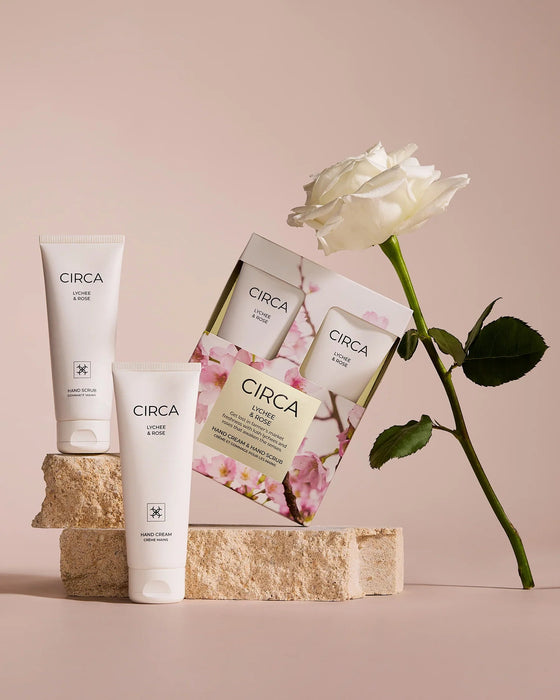 a set of three white cylindrical tubes of CIRCA lotion and rose, along with a box of CIRCA lotion and rose, all set against a light pink background. A single white rose is also present in the scene.