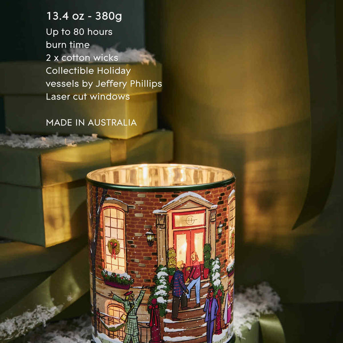 A candle in a decorative container with a winter scene illustration, placed on a surface with a snowy background.