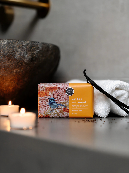 a bar of soap with a bird design, placed on a counter next to a white towel and a pair of black scissors. There are also two lit candles in the background.