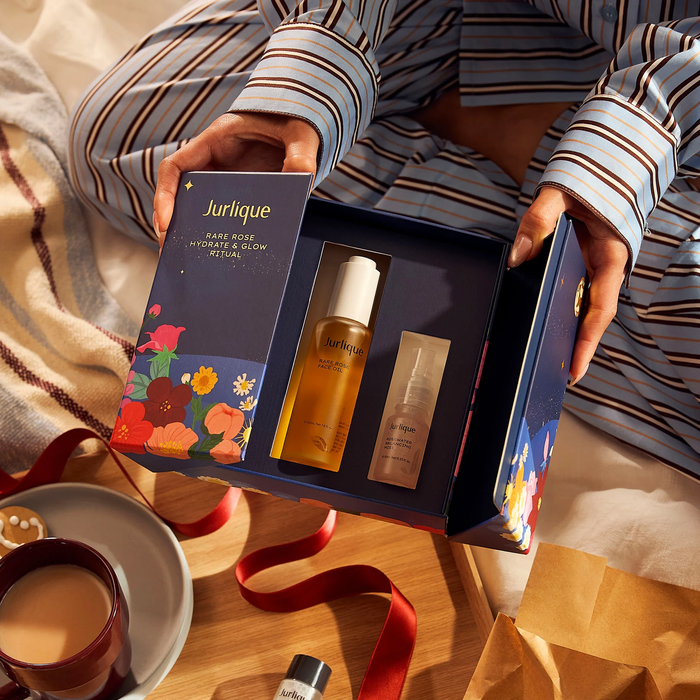 A person is holding a blue box containing a beauty product, with a cup of coffee and a red ribbon visible in the background.