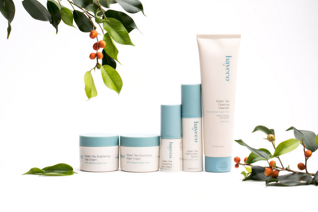 a collection of five skincare products from the brand Bayeco, arranged on a white background. The products include a tube of "Green Tea Cleansing Foaming Night Cream", a jar of "Green Tea Brightening Skin Cream", a jar of "Green Tea Nourishing Eye Cream", a tube of "Green Tea Serum", and a tube of "Green Tea Anti-aging Serum". The products are surrounded by leaves and branches, creating a natural and serene atmosphere.