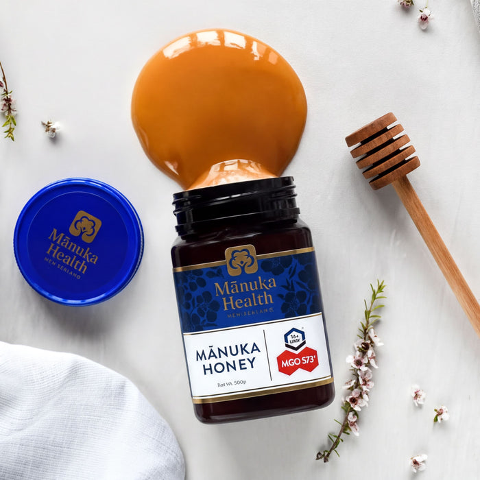 a jar of Manuka Honey, a jar of Manuka Health, and a wooden honey dipper, all arranged on a white surface.