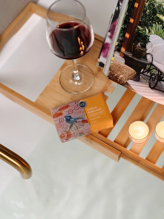 A wooden tray with a glass of red wine, a small box of vanilla and cardamom bath products, and two lit candles, all set on a white surface.