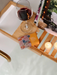A wooden tray with a glass of red wine, a small box of vanilla and cardamom bath products, and two lit candles, all set on a white surface.