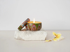 A lit candle in a colorful tin sits on a white rock, with a single yellow flower petal nearby and a small piece of the flower on the floor.