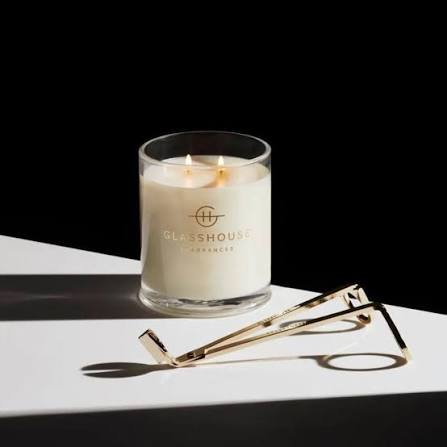 A white candle with the "Glosshouse" logo and three lit candles sits on a white surface, accompanied by a pair of gold-colored candle holders.