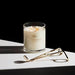 A white candle with the "Glosshouse" logo and three lit candles sits on a white surface, accompanied by a pair of gold-colored candle holders.