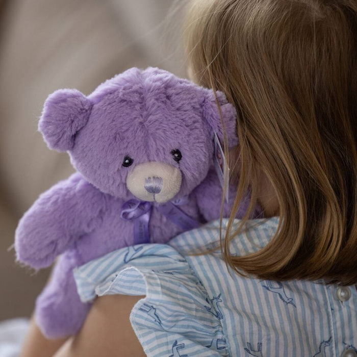 A young girl is holding a purple teddy bear with a blue ribbon around its neck.