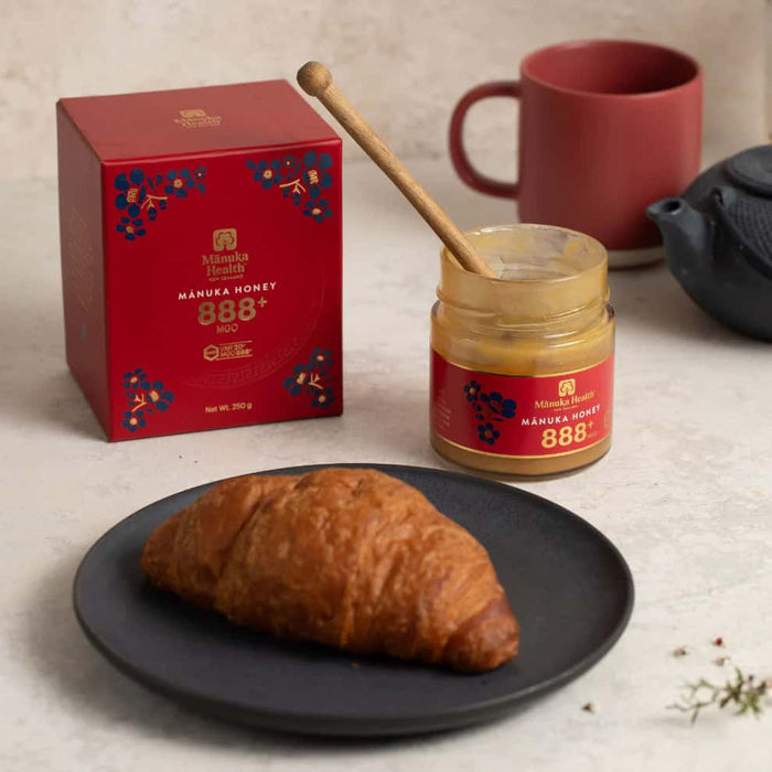 a croissant on a black plate, with a jar of Manuka Honey and a red mug in the background.