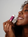 A woman with curly hair is smiling and holding a purple lip balm in her hand.