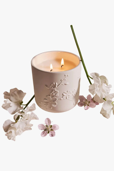 A white ceramic candle holder with a floral design sits on a white background, surrounded by delicate white flowers and a green stem.