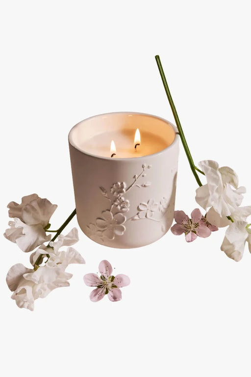A white ceramic candle holder with a floral design sits on a white background, surrounded by delicate white flowers and a green stem.