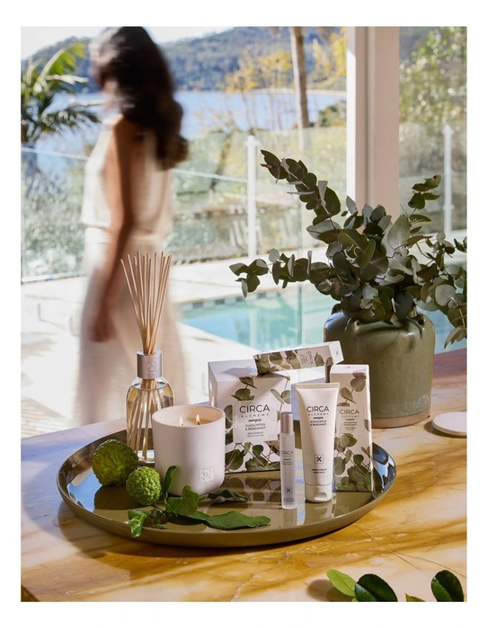 a table with various items, including candles, a vase with greenery, and a tray with a candle and other items. A blurred figure of a woman can be seen in the background, and the setting appears to be a poolside or patio area with a scenic view.