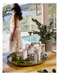a table with various items, including candles, a vase with greenery, and a tray with a candle and other items. A blurred figure of a woman can be seen in the background, and the setting appears to be a poolside or patio area with a scenic view.