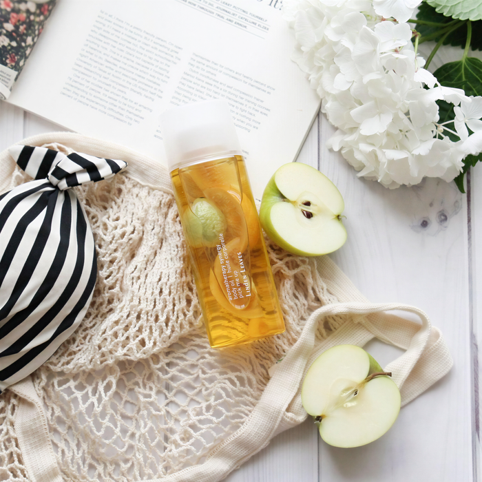 a bottle of liquid, a striped bag, a white book, a white flower, and two apple slices arranged on a white surface.