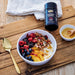 A bowl of granola, yogurt, and fresh fruit, accompanied by a spoon, a bottle of Manuka Honey, and a small bowl of honey on a wooden surface.