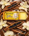 a yellow tin of Lucamar lip balm surrounded by vanilla pods, blackberries, and white orchid flowers.